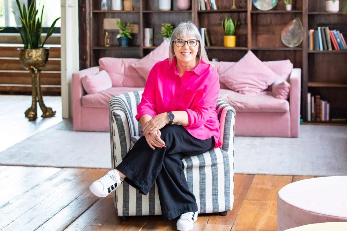 Debbie Green portrait headshot, smiling woman wearing a pink top black pants and white sneakers sitting on striped chair