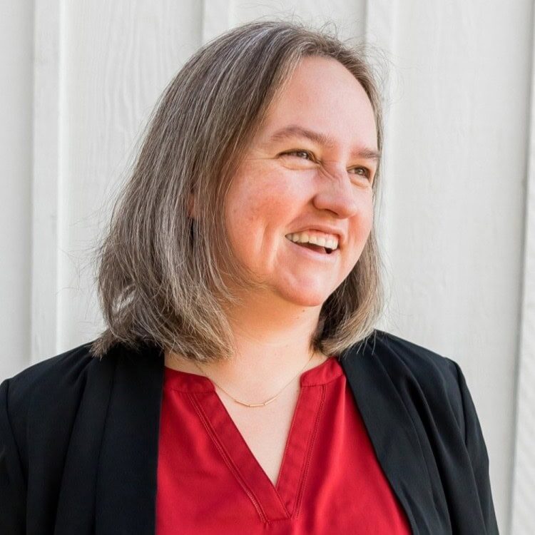 Andrea Fruhling PCC headshot photo, smiling woman with gray hair wearing black blazer and red top in front of white wood wall