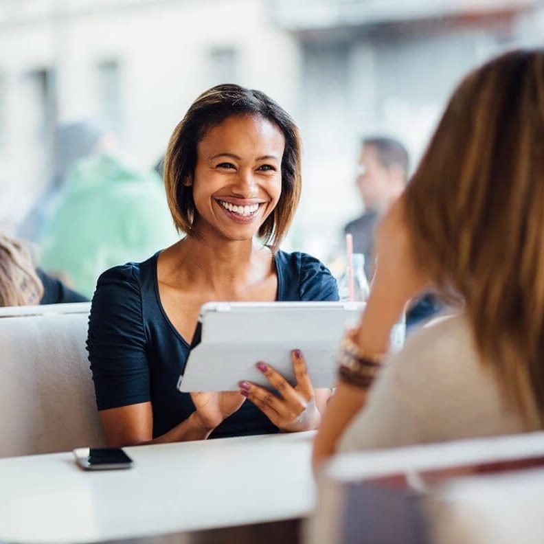 A smiling woman holding a tablet, chatting with another woman at a cafe.