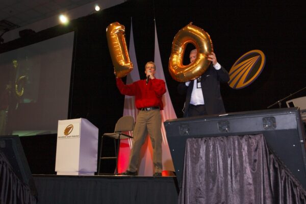 Two men stand on stage holding gold balloons—one displaying the number "1" and the other displaying the number "0"—celebrating the 10th anniversary of the International Coaching Federation at the annual conference.