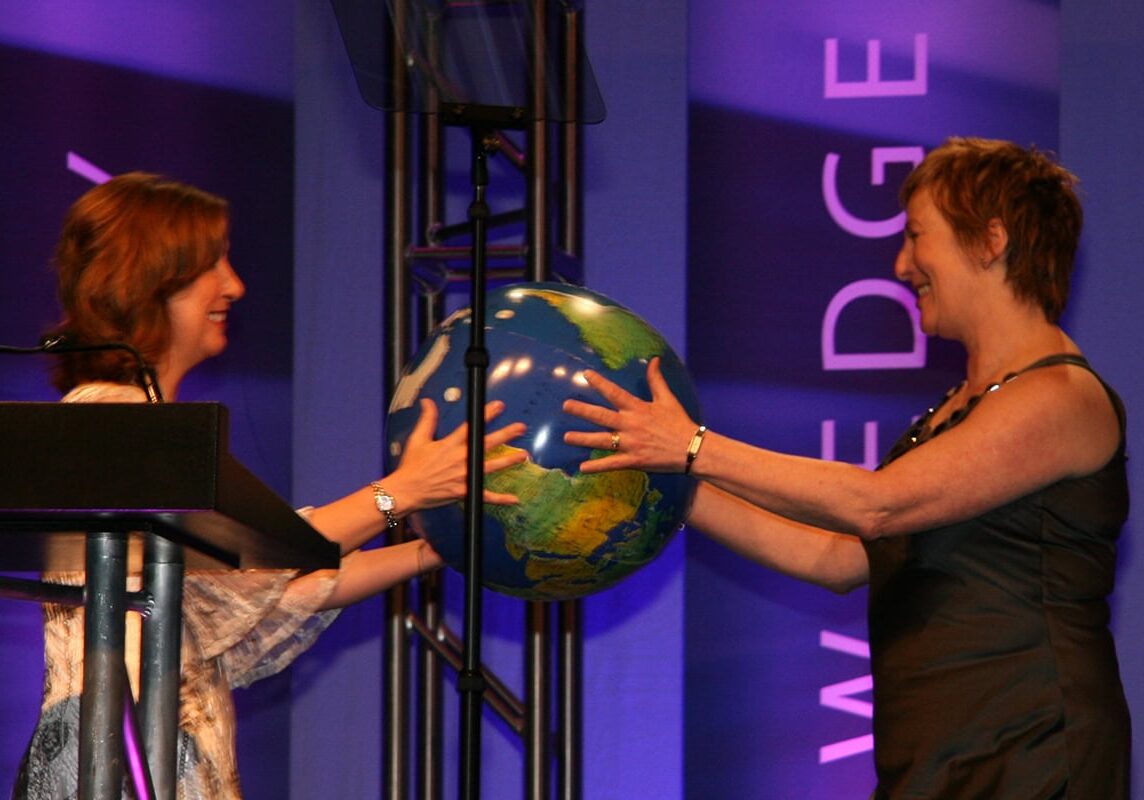A woman passing a globe to another woman, symbolizing the transition of leadership to the 2009 ICF Global Board President.