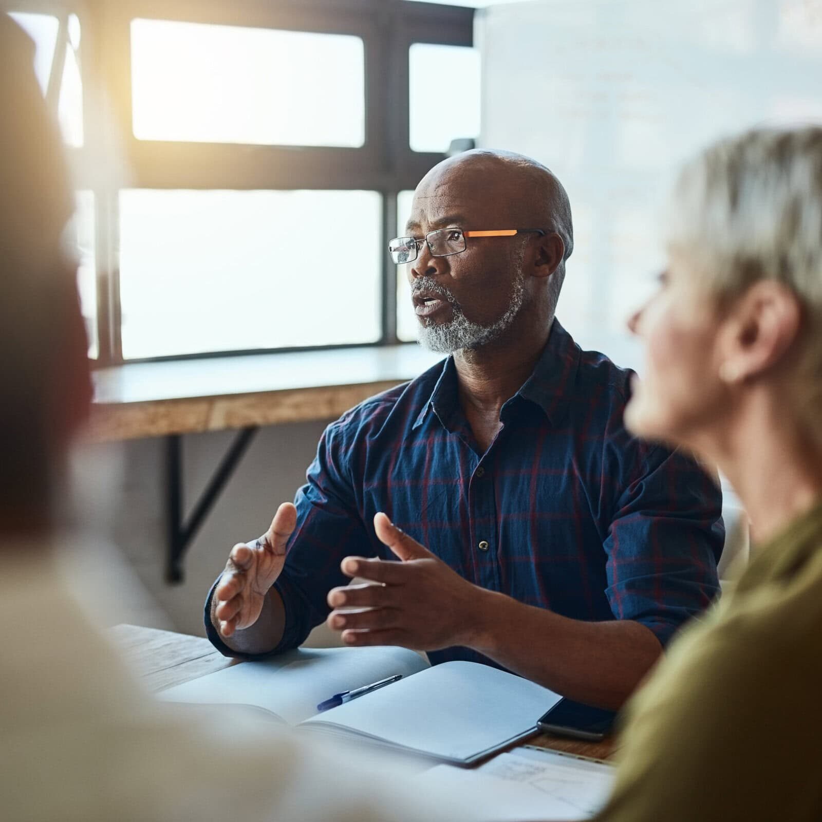 A man holds a meeting in an office and is talking and speaking with the other professionals in the room to collaborate on a project