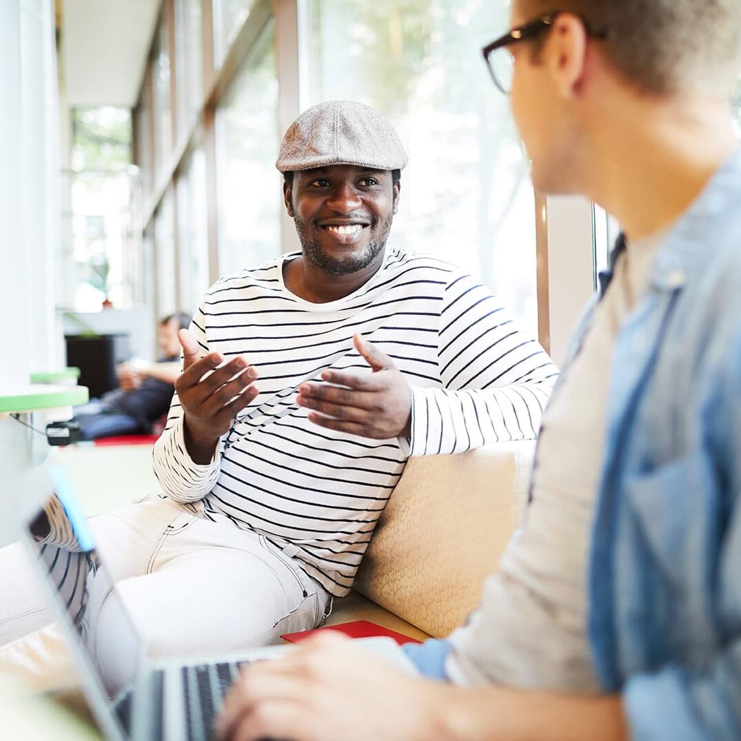 Two men engaged in a lively conversation at a community event, one smiling warmly and gesturing expressively, seated indoors near large windows with natural light filtering in.