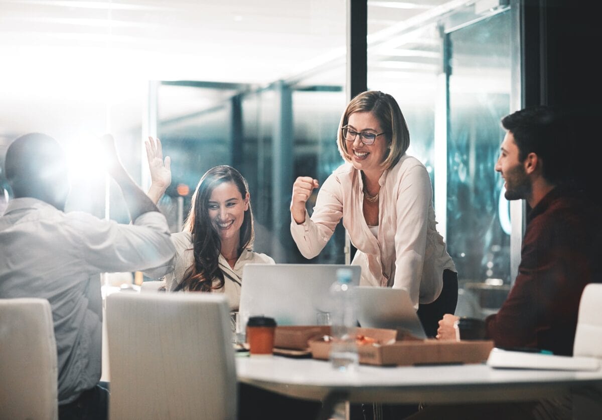 In a bright, modern office, four colleagues sit around a table with laptops and coffee. Two are high-fiving, one is smiling and leaning forward, and another is cheering, showing excitement and teamwork.
