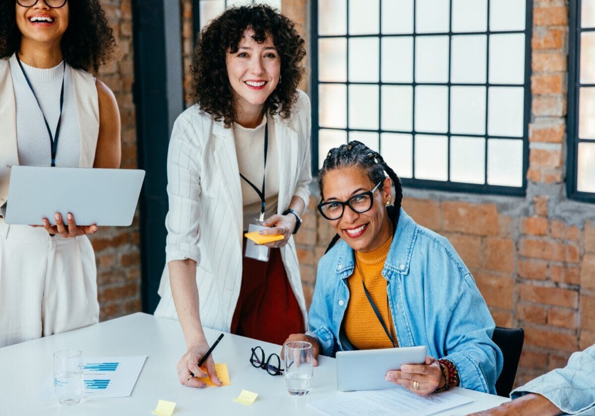 Three female colleagues smiling and collaborating around a table with papers and tablets.