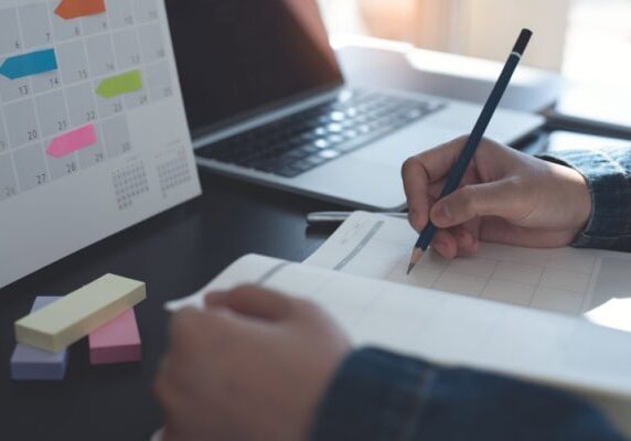 A person writing in a planner with a calendar and laptop in the background representing goal setting in coaching.