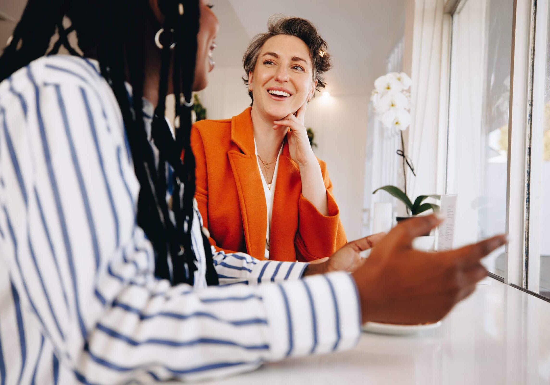 A smiling woman in an orange blazer having a friendly coaching interaction with another woman in a cozy indoor setting.