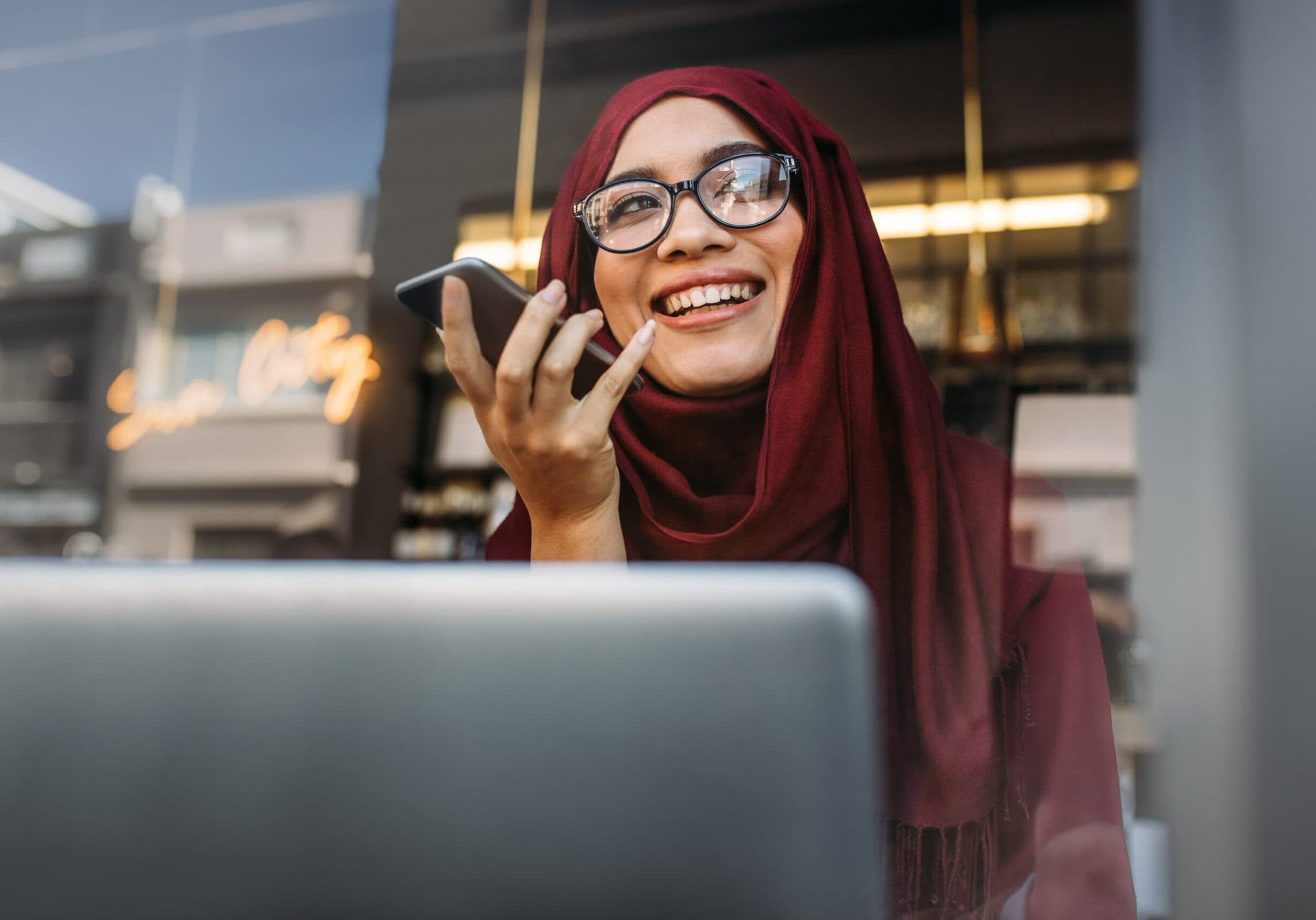 Young professional woman in a maroon headscarf smiling while having a coaching session on her phone in front of a laptop, symbolizing connection and inclusivity within the ICF community.