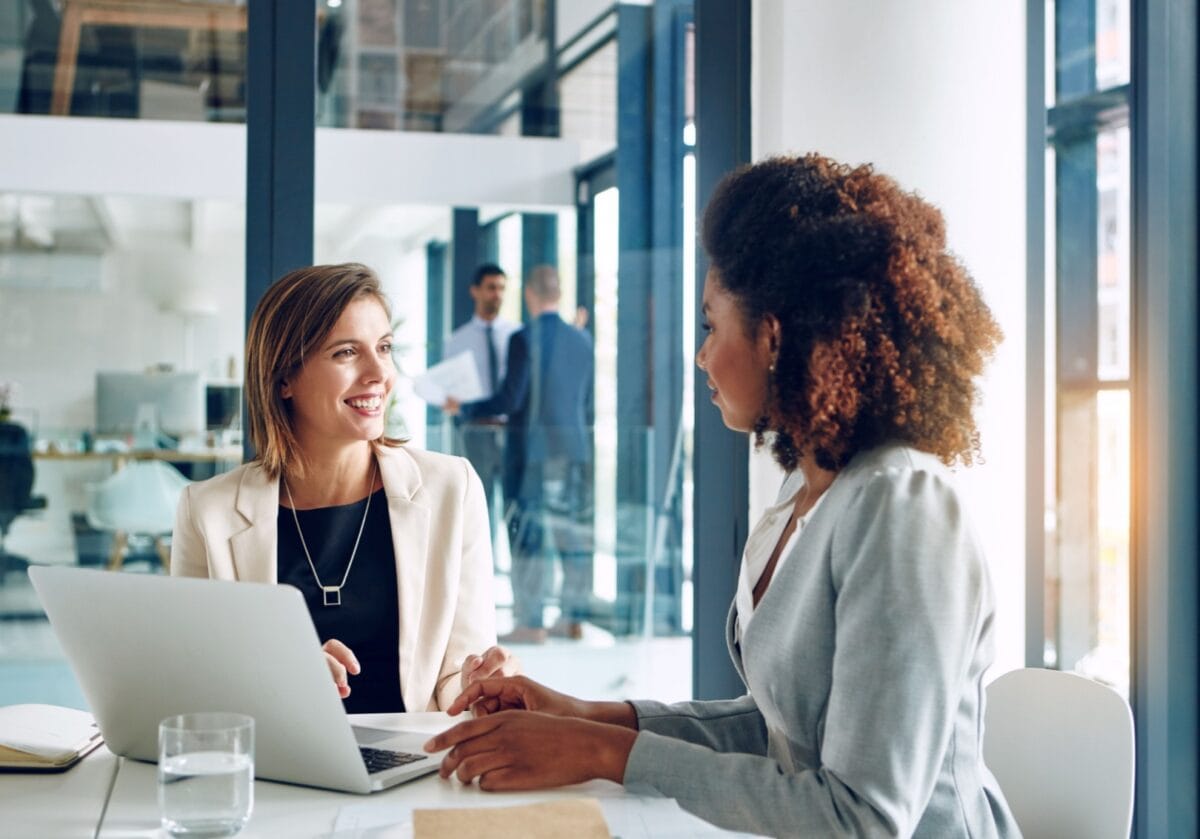 Two diverse young women having a friendly conversation at a table, suggesting one is finding the right coach.