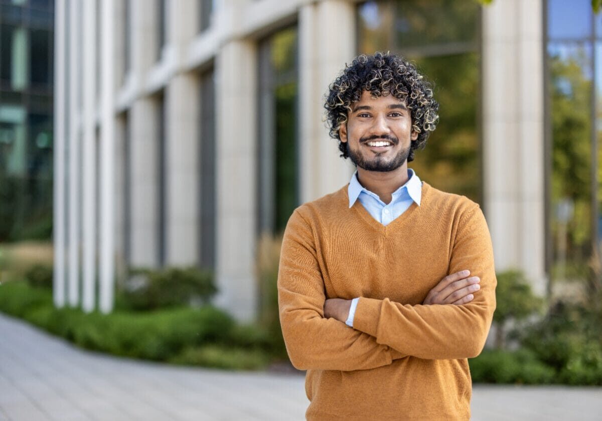 Confident young man with curly hair smiling outside modern office building. Wearing orange sweater with arms crossed, expressing positivity and professionalism.