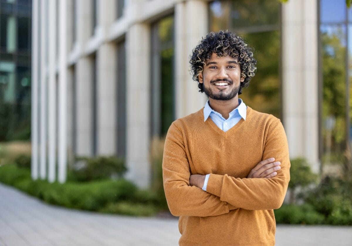 Confident young man with curly hair smiling outside modern office building. Wearing orange sweater with arms crossed, expressing positivity and professionalism.
