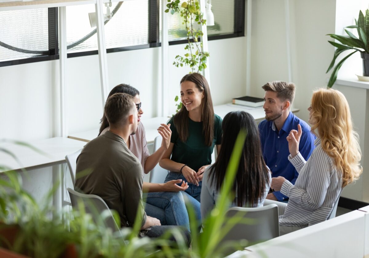 A group of six diverse professionals sits in a circle, smiling and talking in a bright office.
