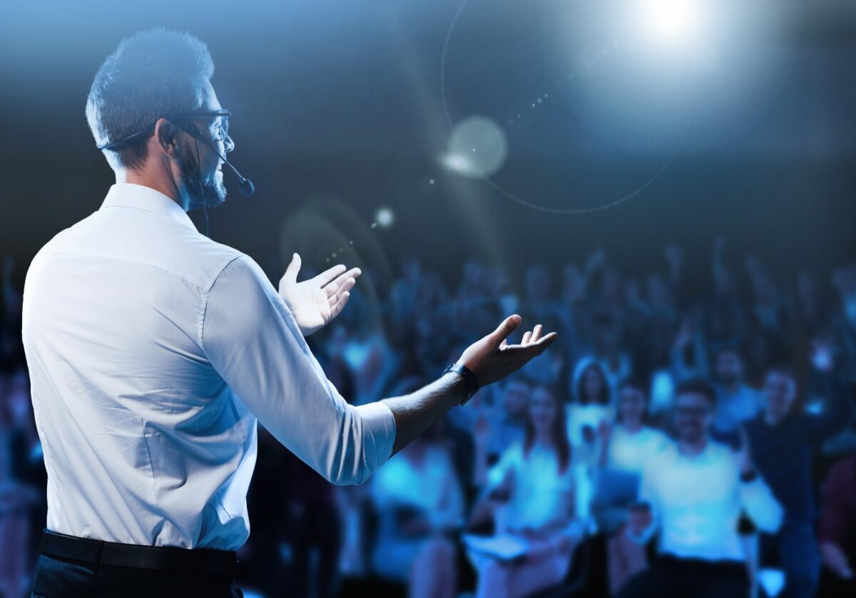 White male motivational speaker with a headset speaks on stage in a dark, crowded room.