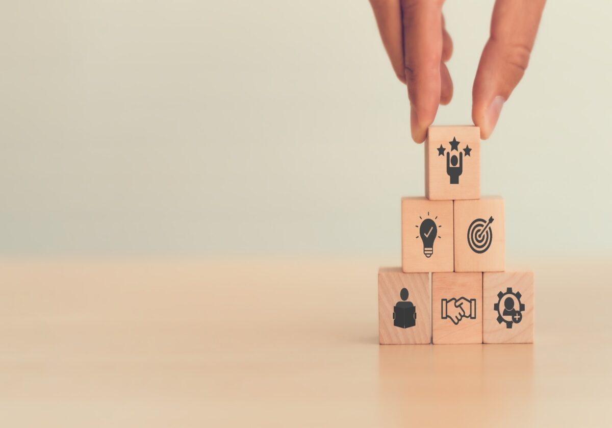 Hand placing a wooden block with three stars on top of a pyramid stack of blocks with business icons.