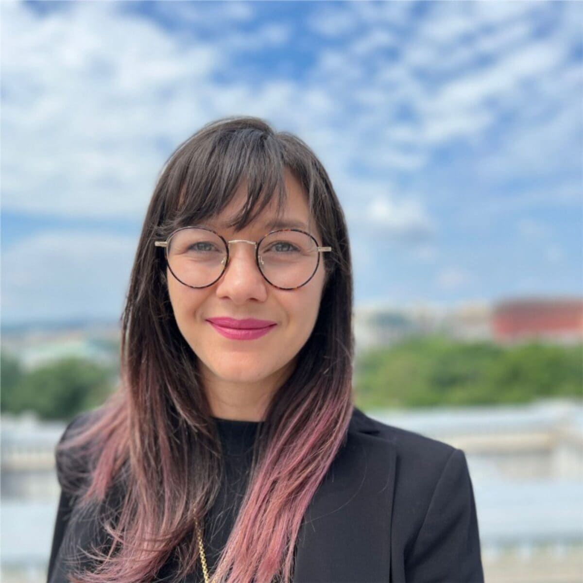 Emily Wenstrom headshot photo, smiling woman with long hair wearing black top and jacket in front of blue sky