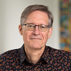 Paul Boehnke PCC ELI MP headshot photo, gray haired man with glasses and dark collared shirt smiling, blurred background